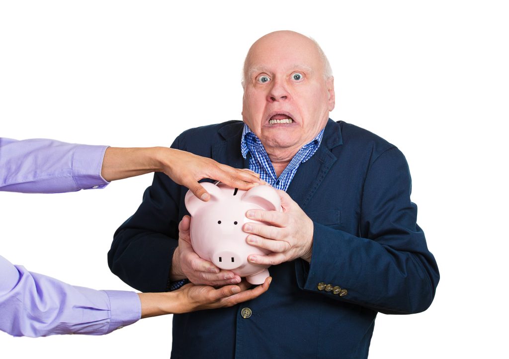 A startled man dealing with elder financial abuse. Elderly man holding piggy bank as greedy scammers hands from out of frame reach in to grab the piggy.