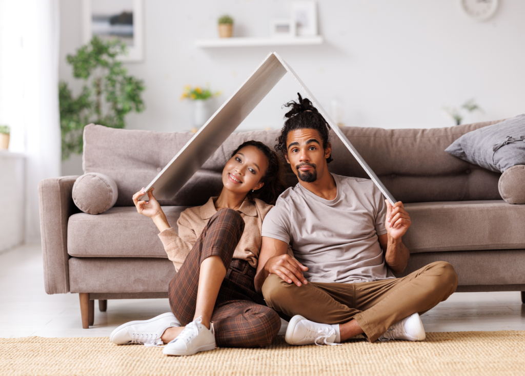 Couple that is search for the best means of Buying a house in 2024. Young man and woman playfully under a roof made of foam board.