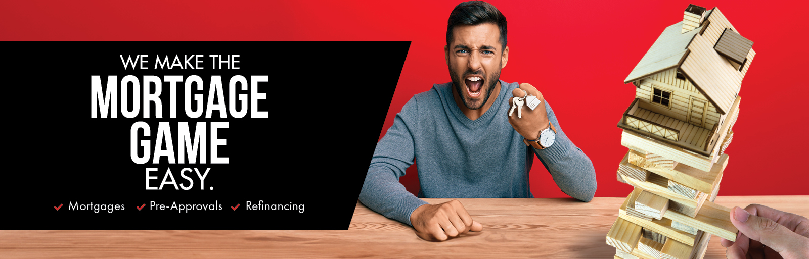 We make the mortgage game look easy. Millenial aged man cheering as he holds house keys, putting together a tumbling block tower house on a table in front of him.